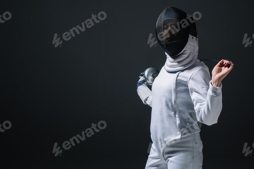 Preview: Fencer in fencing mask looking at camera while holding rapier isolated on black