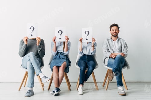 Preview: Handsome man smiling at camera near employees holding cards with question marks while waiting job