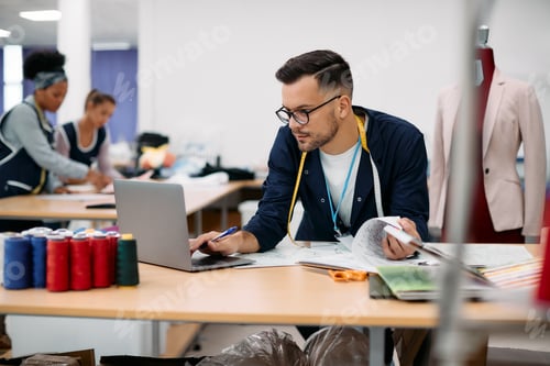 Preview: Young tailor working on laptop at clothing factory.