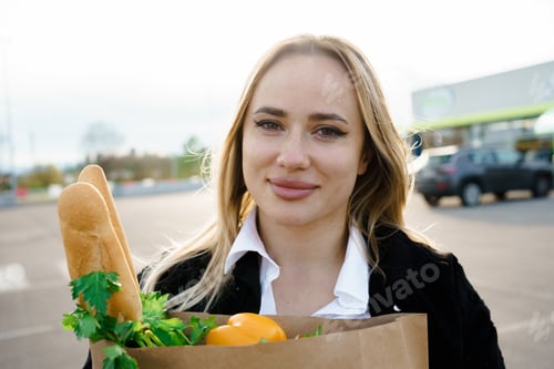 Preview: A close-up portrait of a beaming woman holding a paper bag containing an assortment of fresh and