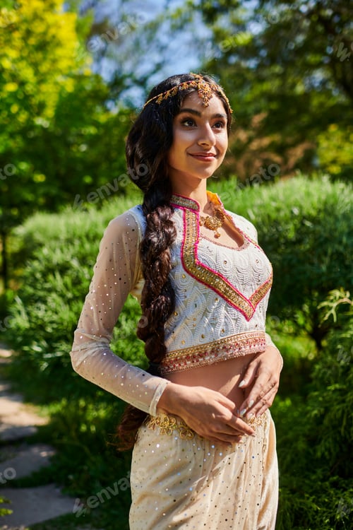 Preview: stylish and young indian woman in trendy traditional outfit standing and posing in park