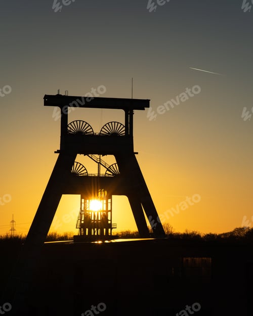 Preview: Bright sun in yellow sunset sky shining behind a mining tower in the field