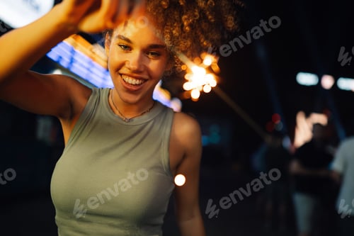 Preview: Happy young woman holding sparkler at night party celebrating with friends