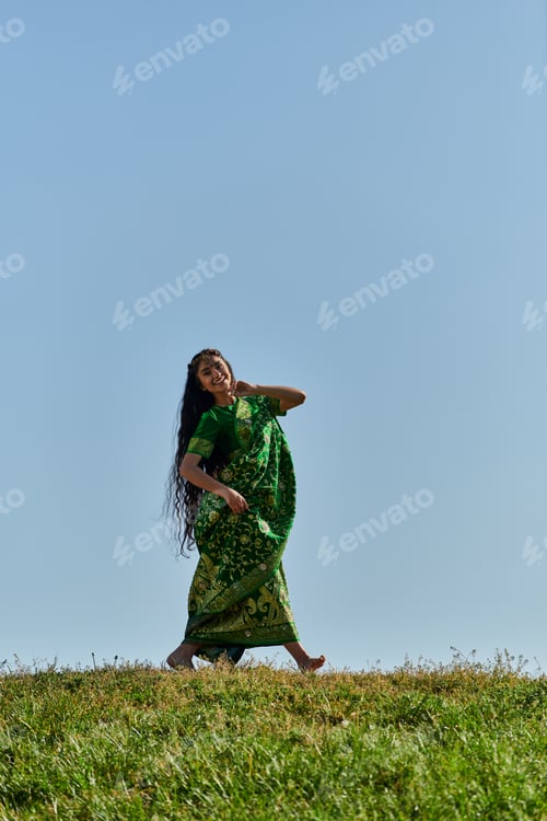 Preview: summer leisure, carefree indian woman in sari walking on green meadow under blue cloudless sky