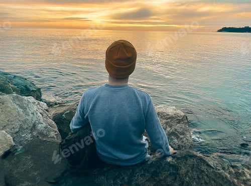 Preview: Young man in casual clothes sits on the rocks near the sea. Caucasian guy in a blue sweater