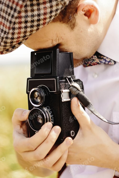 Preview: stylish hipster man looking and holding medium format film camera in sunny sunflower field