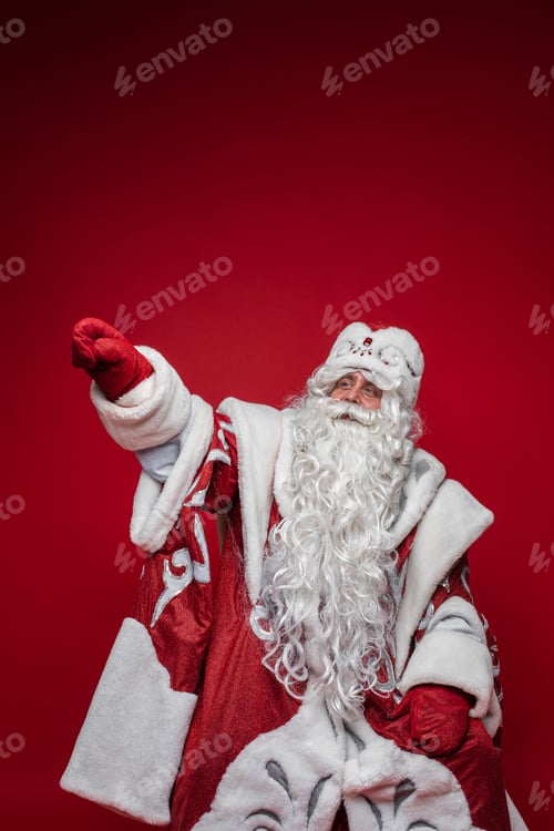 Preview: Senior Santa Claus with gray beard pointing by hand away on red background with copy space