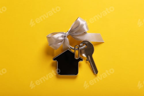 Preview: Key with trinket in shape of house and grey bow on yellow background, top view. Housewarming party