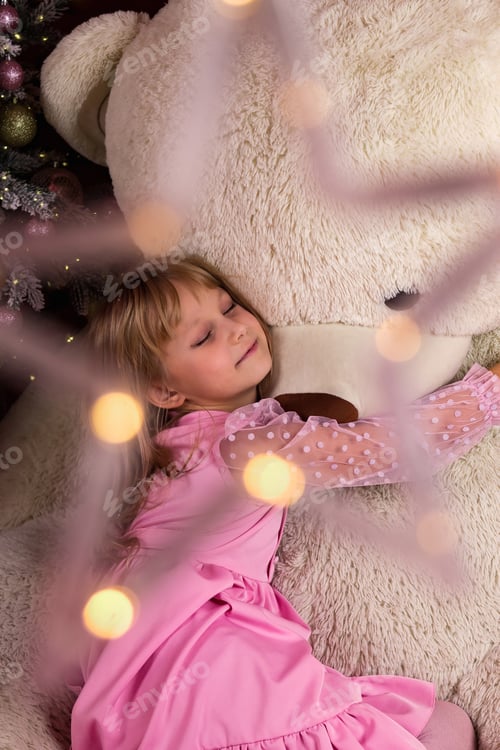 Preview: Charming girl dressed in pink embraces a giant teddy bear in a festive Christmas setting