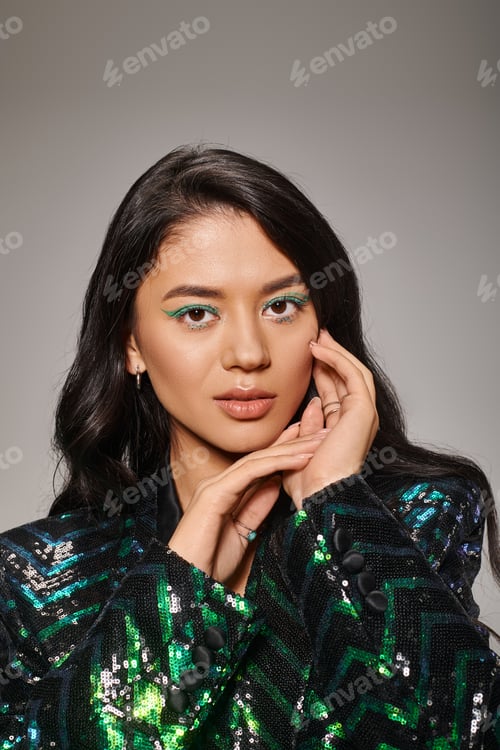 Preview: brunette asian woman in green jacket with sequins and bold eye makeup posing on grey backdrop
