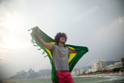 Preview: Smiling young man holding up Brazilian flag on Ipanema beach, Rio De Janeiro, Brazil