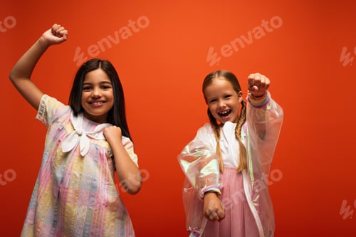 Preview: joyful girls looking at camera and showing clenched fists while having fun isolated on orange