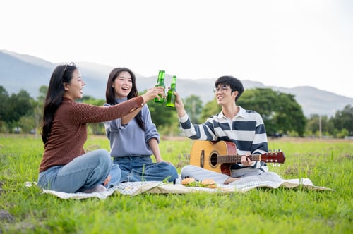 Preview: Asian woman and man friends clinking bottles on picnic mat in grass field with mountain view and sky