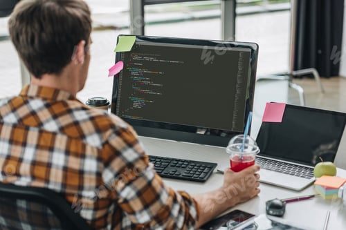 Preview: back view of young programmer holding glass of juice while working on computer in office
