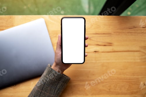 Preview: a woman holding mobile phone with blank white desktop screen with laptop on table