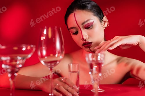 Preview: seductive asian woman with naked shoulders and artistic visage touching glass of water on red