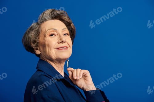 Preview: A stylish woman in her 60s poses confidently, radiating beauty and grace against a blue background.