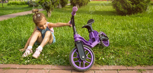 Preview: Cheerful girl sitting resting on the grass in a park near to the purple running bike in summer time.