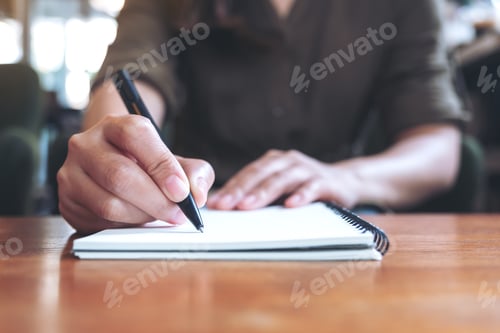 Preview: Closeup image of a woman writing down on a white blank notebook on wooden table