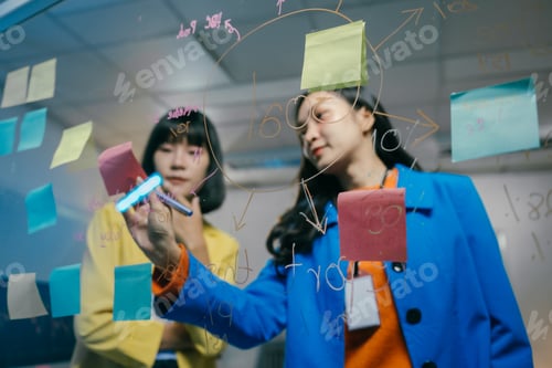 Preview: Two asian businesswomen are brainstorming and using colorful sticky notes on a glass wall to visuali