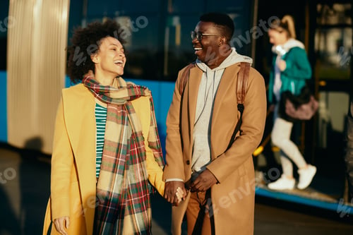 Preview: Cheerful black couple holding hands and having fun at bus station.
