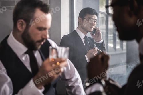 Preview: Serious asian businessman smoking cigar and talking on smartphone while colleagues drinking whisky,