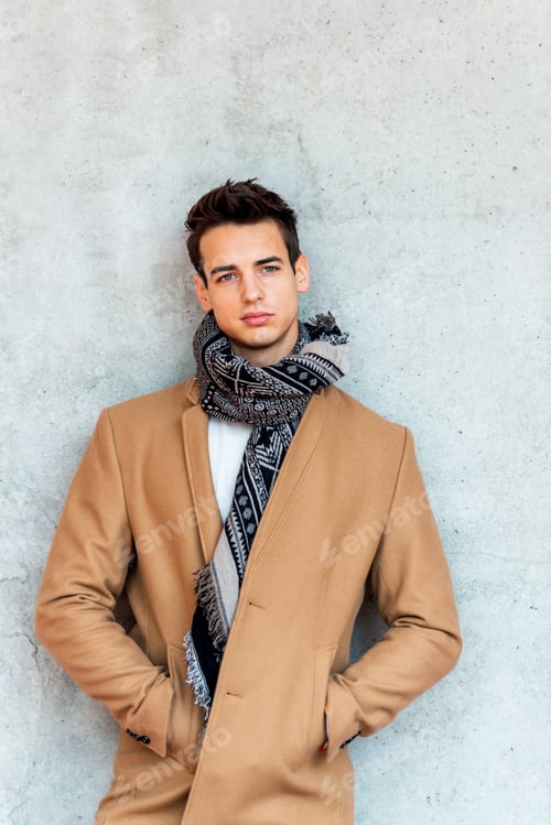Preview: Front view of trendy young man wearing coat and scarf leaning on a wall while looking camera