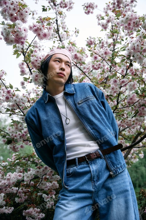Preview: Young man in stylish denim outfit enjoying a spring day in a floral park setting