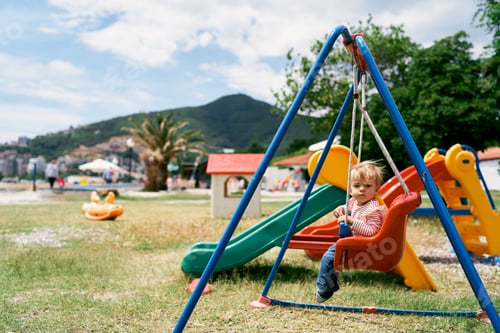 Preview: Small kid sits on a colorful swing on the playground