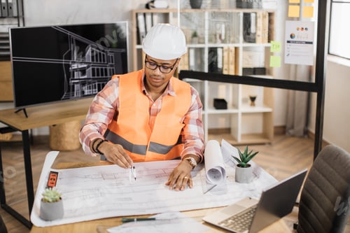Preview: Young african builder, engineer browsing in laptop and looking through papers or sketches.
