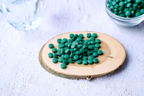 Preview: Green round spirulina pills on natural wood plate on a simple minimalistic gray concrete background.