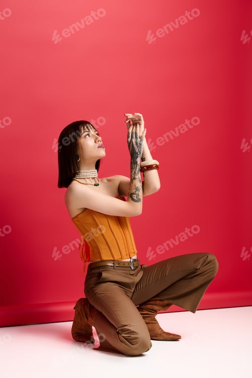 Preview: Charming young woman in stylish attire kneels gracefully against a vibrant red backdrop