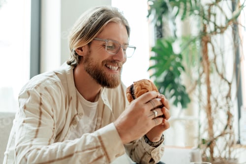 Preview: Happy young man with beard and glasses enjoying eating fresh croissant