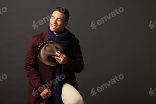 Preview: smiling mixed race man in scarf holding hat with closed eyes on black background
