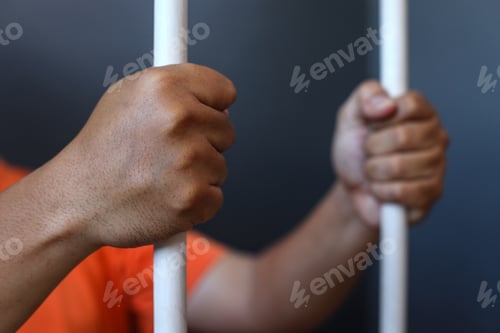 Preview: Close up of hands of prisoner in orange shirt inside the bars of a prison