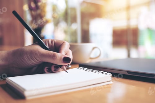 Preview: woman's hand writing on a blank notebook with laptop , tablet and coffee cup on wooden table