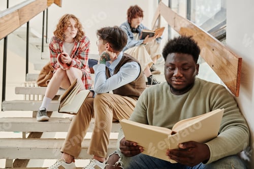 Preview: Serious African American teenager in casualwear reading book against classmates
