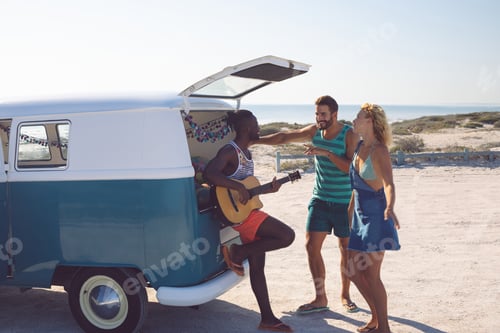 Preview: Side view of happy group of diverse friends having fun together near camper van at beach