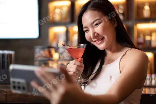 Preview: Beautiful woman is holding cocktail and using smartphone to selfie during having enjoy to relaxing