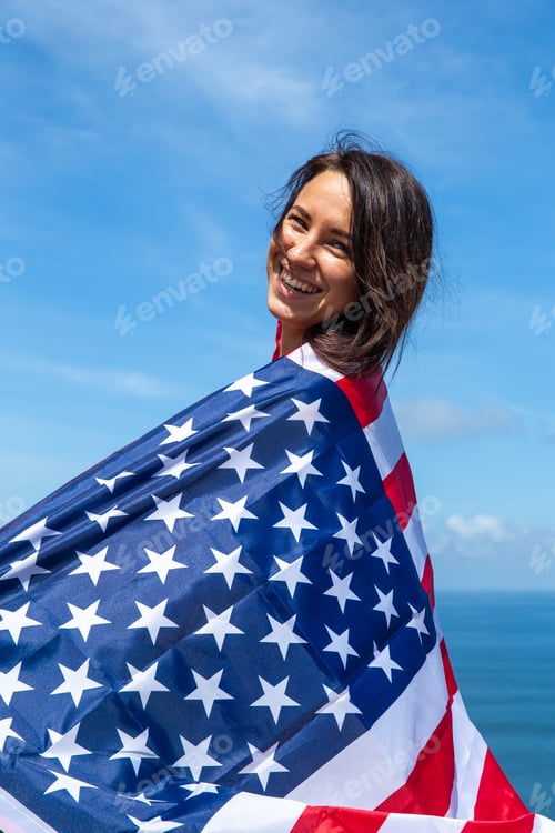 Preview: Beautiful woman is holding United States of America flag and standing against blue sky