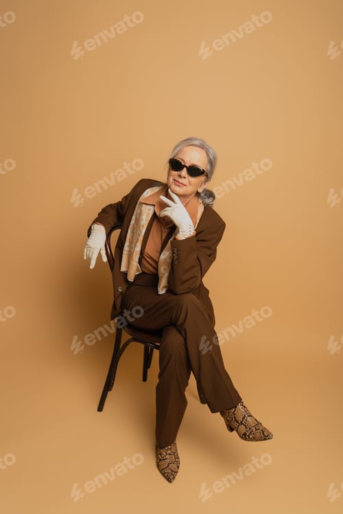 Preview: full length of smiling elderly woman in brown suit and sunglasses sitting on chair and looking at