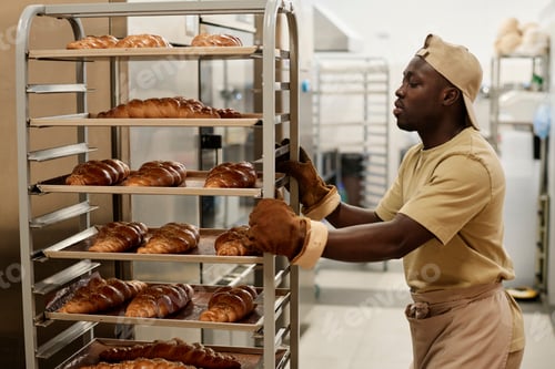 Preview: Black young man moving shelves with freshly baked breads in bakery kitchen