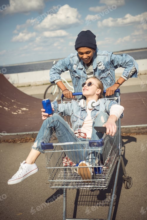 Preview: Boy carrying happy girl with can in shopping cart near ramp at skateboard park