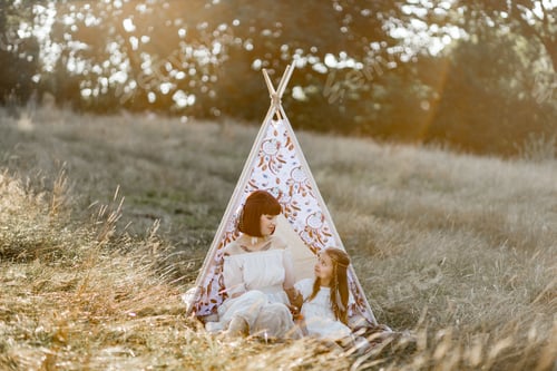 Preview: Mom and little girl wearing white dress, boho feather heair accessories and sitting near teepee tent