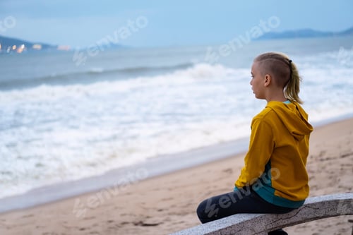 Preview: Lonely teenager looking to sea sitting on the waterfront. Rear view of teenager against seascape.