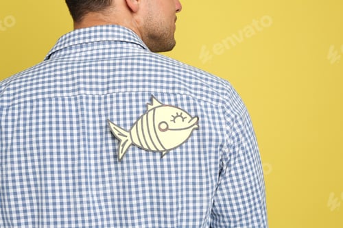 Preview: Man with paper fish on back against yellow background, closeup. April fool's day