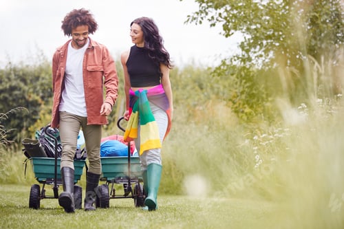 Preview: Young Couple Pulling Trolley Filled With Camping Equipment Through Field To Music Festival