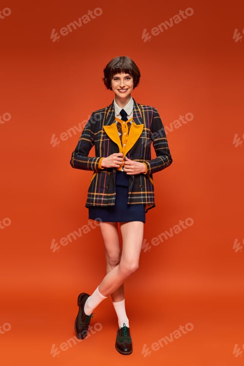 Preview: full length of stylish student in checkered blazer and skirt on orange background, college uniform