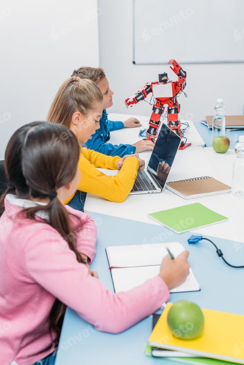 Preview: children writing in notebooks, using laptop and looking at red robot at desk in stem class