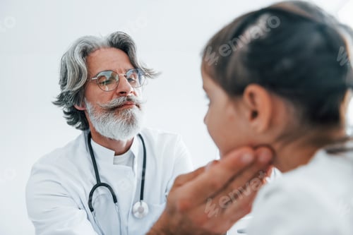 Preview: Senior otorhinolaryngologist with grey hair and beard in white coat is indoors in clinic